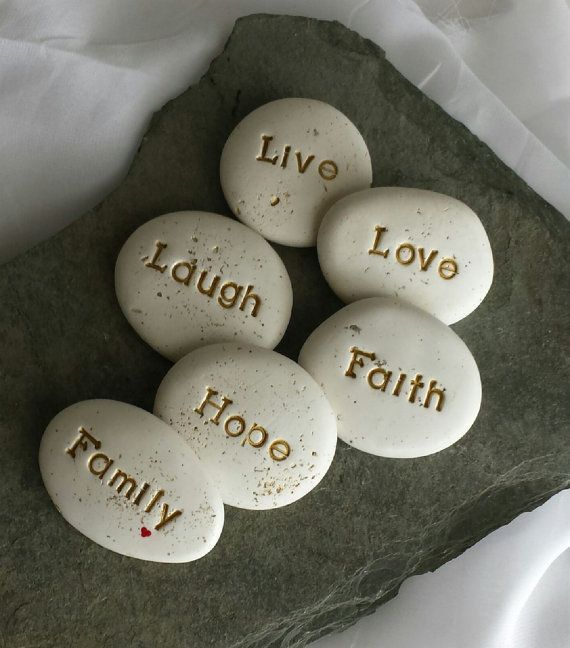 Six smooth white stones arranged on a gray slate surface, each engraved with an inspirational word: Live, Love, Laugh, Faith, Hope, and Family.