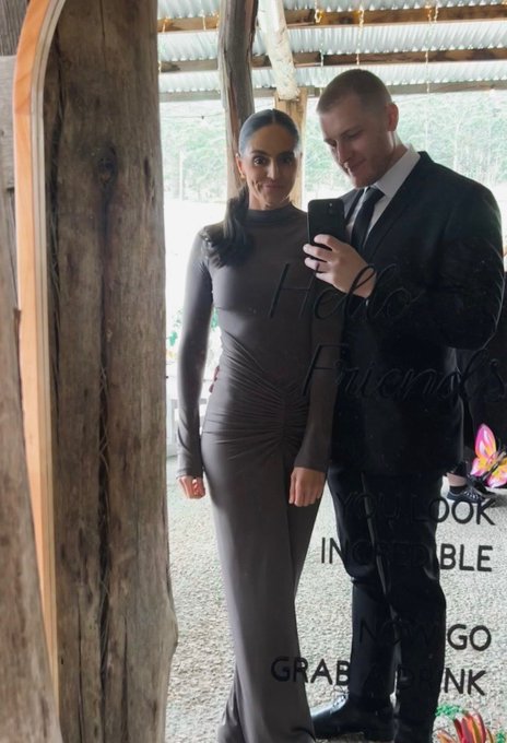 A man and woman in formal attire pose for a mirror selfie in a rustic wooden structure; the mirror displays the etched words 'Hello Friends', 'YOU LOOK INCREDIBLE', 'NOW GO GRAB A DRINK'.