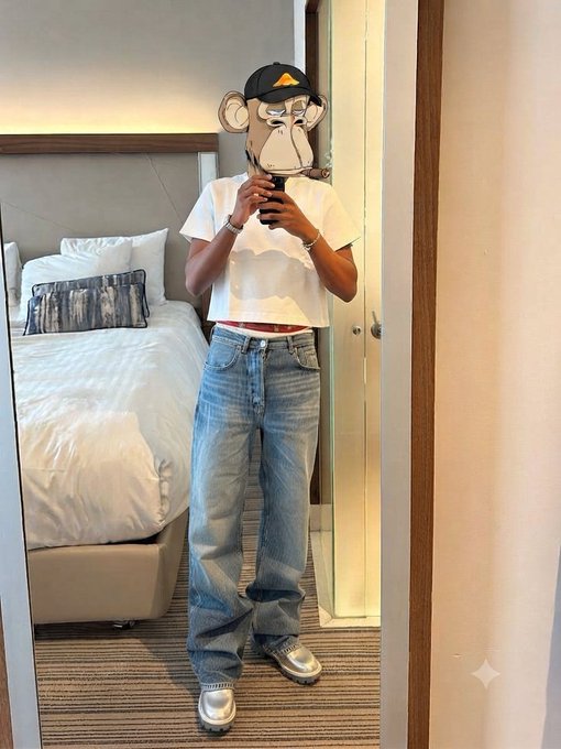 A mirror selfie of a person wearing baggy light blue jeans, a white t-shirt, silver sneakers, and a black cap, with a cartoon monkey face sticker covering their face, standing in a hotel room with a bed visible behind them.