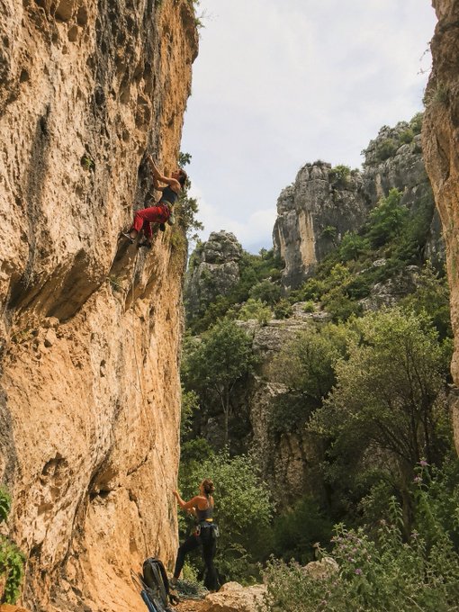 A rock climber scales a steep limestone cliff in a canyon while a belayer stands below, with rocky peaks and green trees in the background.