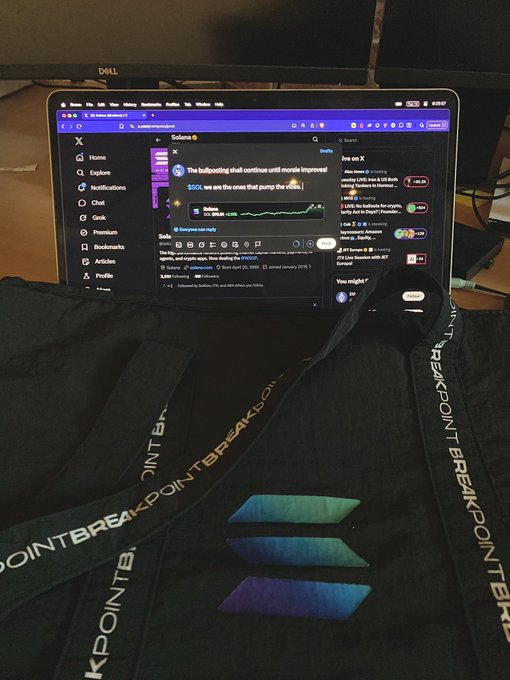A first-person view of someone using a MacBook displaying the X (Twitter) feed, with their black Solana Breakpoint hoodie featuring a holographic logo visible in the foreground.