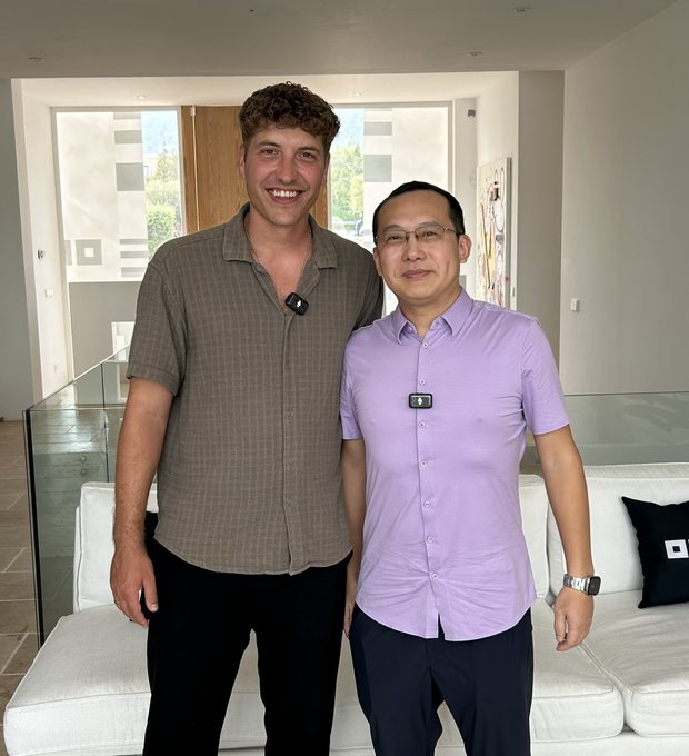 Two men pose together in a modern, well-lit living room. The taller man on the left wears a brown short-sleeved button-up shirt, while the shorter man on the right wears a purple button-up shirt and a wristwatch; both have small microphones clipped to their shirts, suggesting a recorded interview or content creation setup that aligns with the post about speaking with the CEO of OKX, @star_okx.