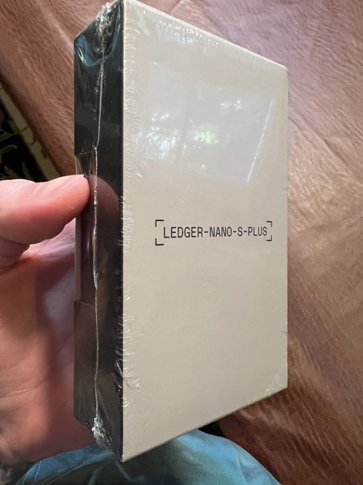 A hand holding a shrink-wrapped white box labeled "LEDGER NANO S PLUS," a hardware wallet device.