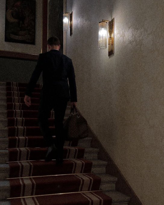 A silhouette of a man in a dark suit carrying a duffel bag as he climbs a dimly lit carpeted staircase with crystal wall sconces.
