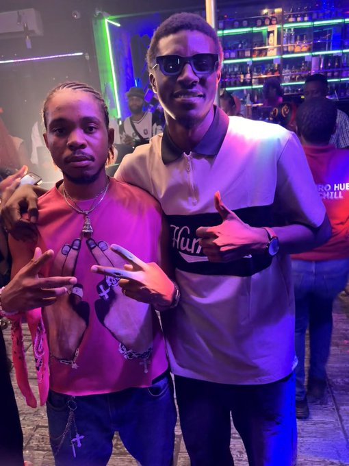 Two young men posing for a photo in a nightclub/bar setting with neon lighting and liquor bottles in the background; one wears a pink shirt with a cross necklace and the other wears a Hurly jersey with sunglasses.