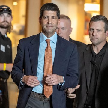 A well-dressed man in a navy suit and orange tie walks through a corridor flanked by security personnel. The image appears to show a Federal Reserve official, likely related to the post about Fed Chair Kevin Warsh sharing a Swan referral code with central bankers at Jackson Hole.