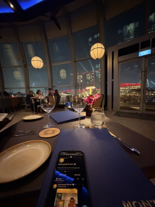 A restaurant table at night with a view of city lights through large windows, set with wine glasses, plates, and a smartphone displaying social media.
