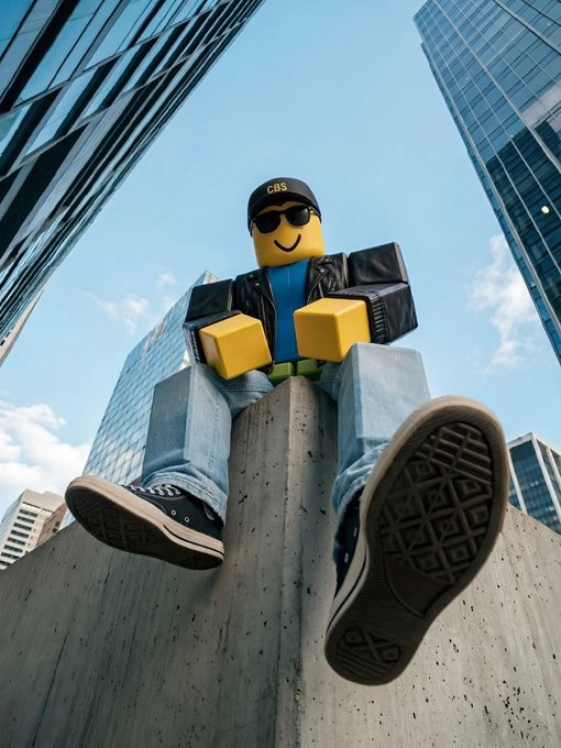 A low-angle photograph of a person dressed as a giant Lego character wearing a CBS cap and sunglasses, sitting on a concrete pillar with skyscrapers in the background.