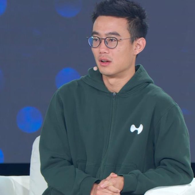 The image features a man wearing glasses, sitting down with his hands clasped together. He is dressed in a green hoodie that has white letters on it. The background of the image shows a blue screen with white circles scattered across it. There are also some words written on this screen, which appear to be related to the post "Hyperliquid supply shock incoming."