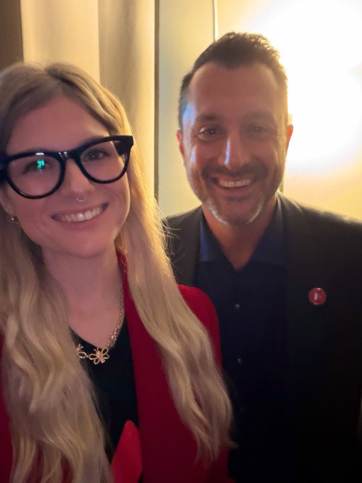 A selfie of a smiling blonde woman wearing black cat-eye glasses and a red jacket, standing next to a smiling man with short dark hair and a beard wearing a black jacket and shirt.