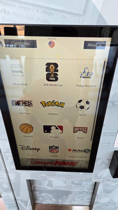 A photo of a digital kiosk screen displaying a menu with a US flag icon at the top and category icons for ShopALL, FIFA World Cup, Pudy Penguins, One Piece, Pokémon, Soccer, Basketball, Bicycle, Disney, Football, and Magic: The Gathering, with partially visible red text at the bottom.