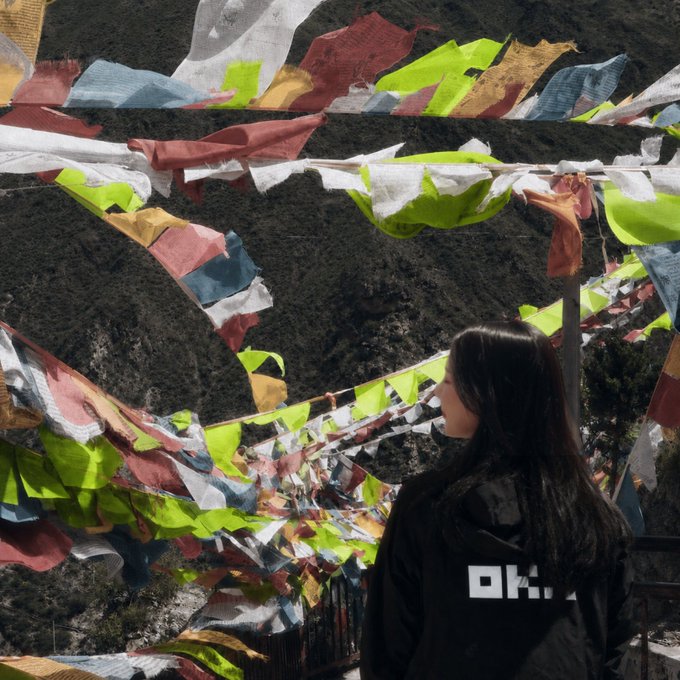A woman with long dark hair wearing a black jacket with 'OKX' on the back looks out at strings of colorful Tibetan prayer flags blowing in the wind against a mountainous backdrop.