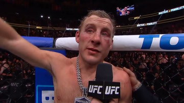 UFC fighter Stipe Miocic, shirtless and visibly bruised, raises his arm in victory during a post-fight interview inside the octagon.