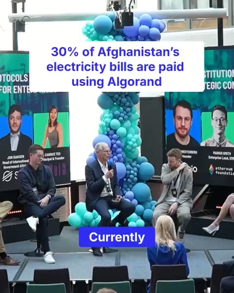 Three men sit on stage at what appears to be a blockchain conference, seated in front of a backdrop featuring balloon decorations and screens displaying other speakers' names. Text overlaid on the image reads "30% of Afghanistan's electricity bills are paid using Algorand," highlighting Algorand's real-world utility in the country.