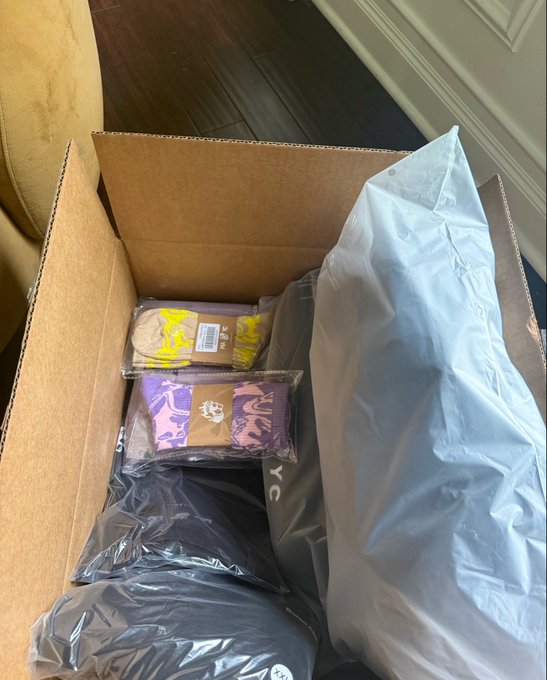 The image shows a cardboard box filled with various items including clothing and accessories such as socks and hats. The box appears to be opened or partially open, revealing its contents. There are at least six pairs of socks visible inside the box, along with two hats. A person wearing a black shirt can also be seen in the background, possibly observing the contents of the box.