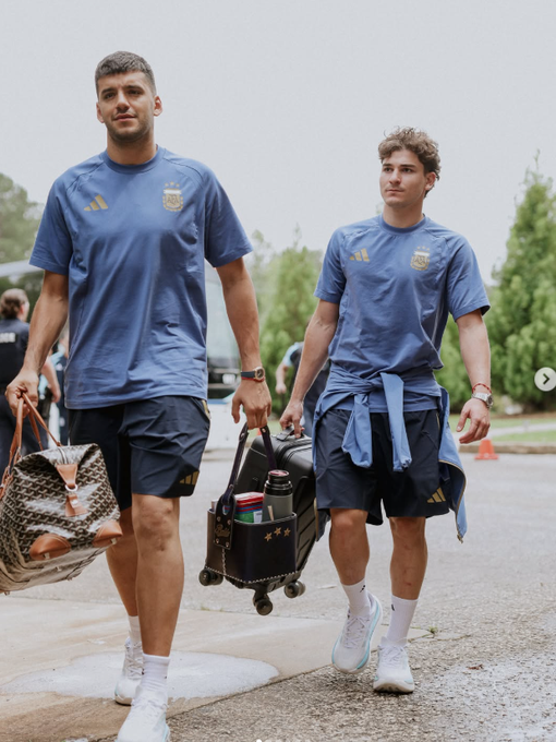 The image shows two tall men in matching blue Argentina national team athletic shirts and dark shorts walking outdoors, carrying luggage including a monogrammed duffel bag and a rolling suitcase. The image does not appear to relate to the post's content about Web3 infrastructure and developer experience with @NucleusCodes, as it depicts athletes rather than anything related to blockchain technology or software development.
