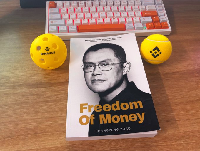 The book "Freedom Of Money" by Changpeng Zhao (CZ) sits on a wooden desk alongside a red and white mechanical keyboard and two yellow Binance-branded items (a wiffle ball and a tennis ball).