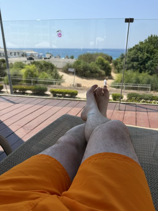 A first-person perspective photo showing a person's legs in orange shorts resting on a lounge chair overlooking a coastal scene with the ocean and beach visible through a glass railing.