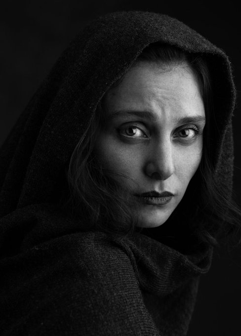 A black-and-white portrait of a woman wrapped in a hooded shawl, her face illuminated against the dark background with an intense, contemplative gaze. The image evokes a sense of mystery and solemnity, aligning with the post's reference to a Tezos-based NFT artwork titled "Liya."