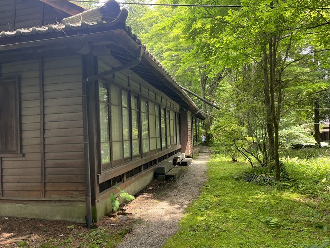 A traditional wooden Japanese building with a tiled roof, large windows, and a gravel path beside it, surrounded by lush green trees and mossy ground.