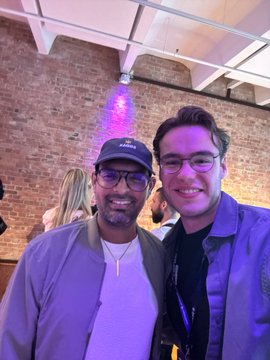 The image shows two men standing next to each other with smiles on their faces, posing for a picture at an event. One of them has a purple jacket on while the other one wears glasses. They are both wearing ties, which adds to their formal appearance. The background features a brick wall and a ceiling light that illuminates the scene. There is also another person visible in the image who appears to be part of the same gathering or event.