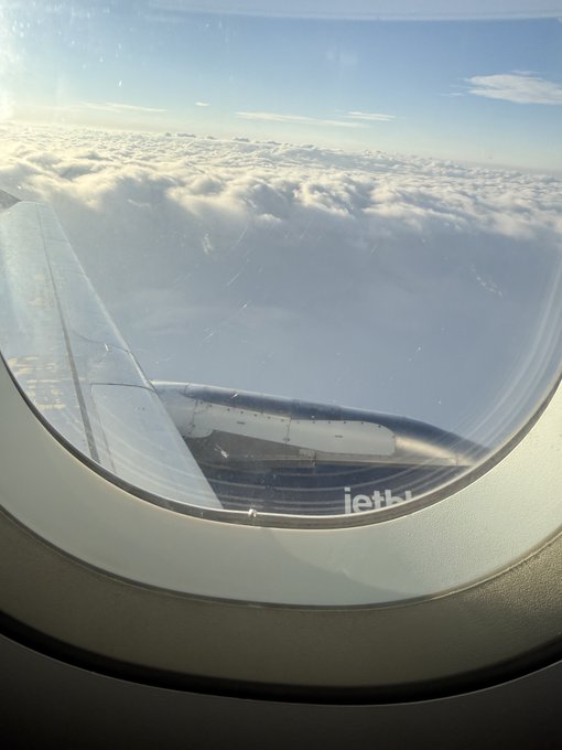 View through an airplane window showing the wing, clouds below, and blue sky, with reversed 'United' text visible on the fuselage.