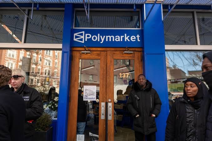 The image shows people outside a storefront with a prominent blue-and-white “Polymarket” sign above the entrance and the street number “137” on the door. It illustrates the post’s report that Polymarket is among prediction-market platforms continuing to offer sports betting despite cease-and-desist letters from Ohio regulators.