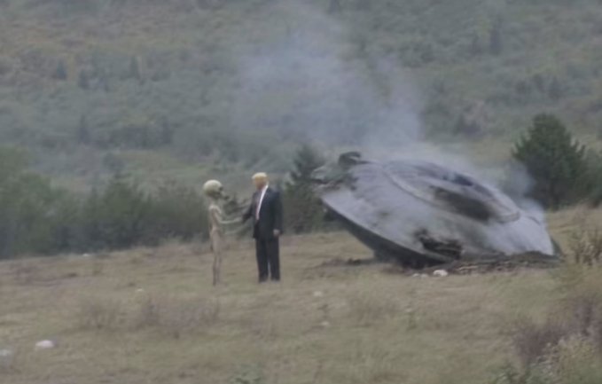 A staged or edited image showing a man in a suit shaking hands with a grey alien in a grassy field with a crashed flying saucer in the background.