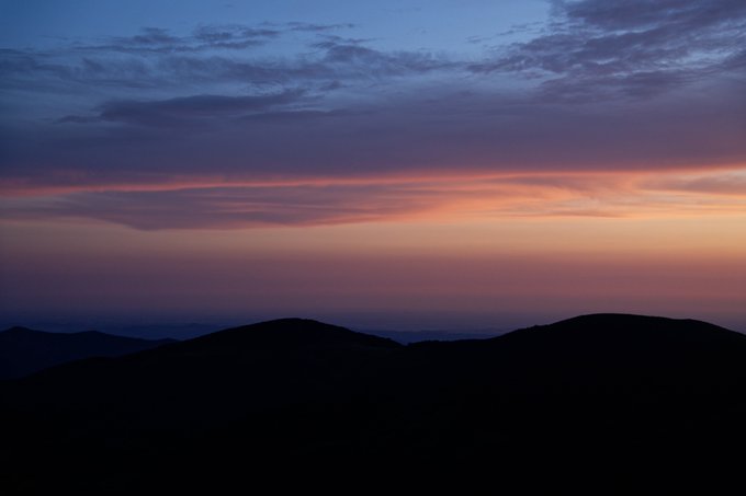 A serene landscape photograph showing silhouettes of mountain ridges against a sunset sky with pink, orange, and blue gradient clouds.
