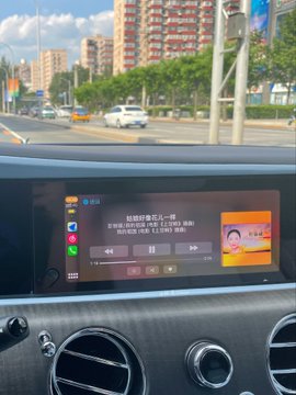 The image captures a driver's perspective from behind the wheel of a car on a city street, with the infotainment screen displaying Chinese text about earning good days through hard work and enjoying the holiday—tying into the National Day message about rest and self-care after diligent effort. Tall buildings and traffic are visible through the windshield, suggesting a quiet moment of relaxation behind the wheel.