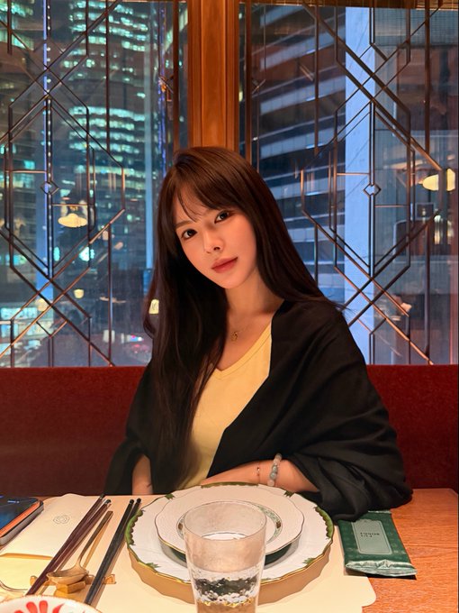 A young woman with long dark hair sits at an elegant restaurant table with an empty plate and water glass before her, dressed in a black blazer over a yellow top, with a nighttime city skyline visible through the geometric-patterned window behind her. The caption "不是薄肌女孩 是抄底女孩" (Not a slim girl, but a "bottom-fishing" girl) plays on the empty plate in front of her, humorously suggesting she's a savvy investor "buying the dip" rather than a delicate eater awaiting her meal.