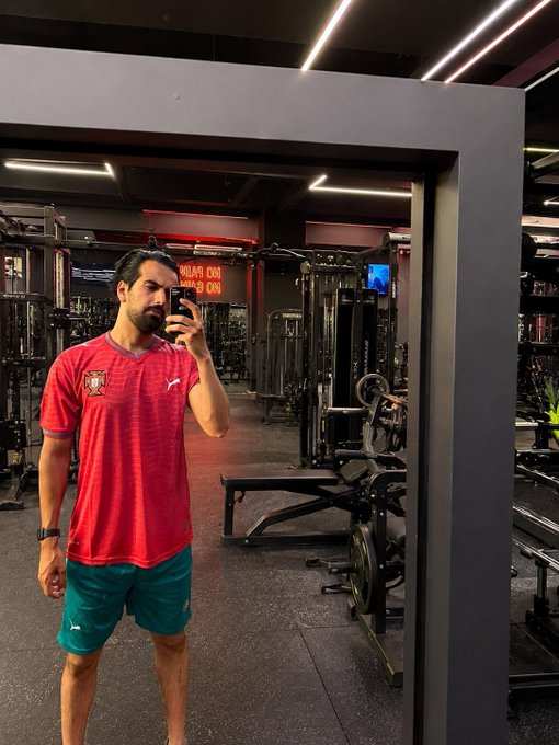 A man with a beard taking a mirror selfie in a gym, wearing a red Portugal national football jersey and green Puma shorts, with weight machines and a 'NO COACH NO GAINS' neon sign visible in the background.