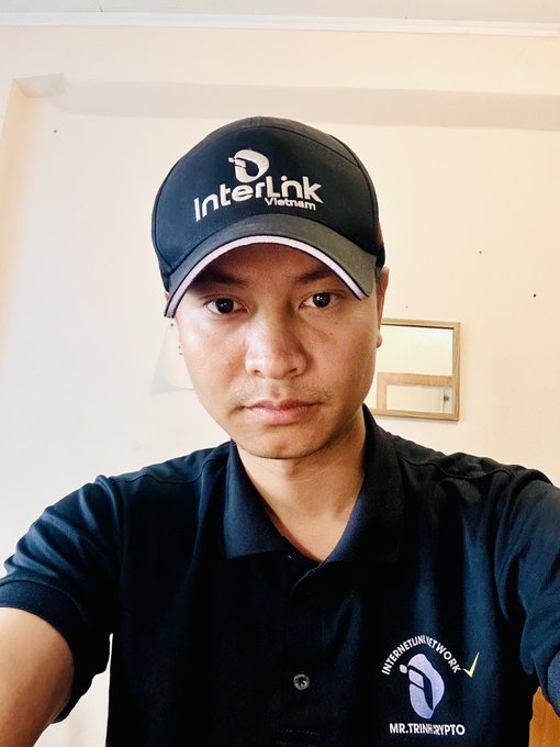 A selfie of a man wearing a dark 'InterLink Vietnam' cap and a matching 'Internet Network' polo shirt, standing indoors against a white wall.