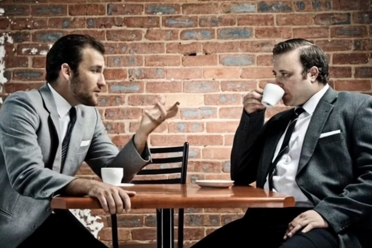 The image shows two men sitting at a table, engaged in conversation while drinking coffee from cups. One of them has a tie visible, indicating that they are dressed for a formal occasion or work setting. The man with the tie appears to be reading something on his phone, possibly checking an email or message. The post reads: "CARDANO IS DEAD", which seems unrelated to the scene in front of it and does not provide any context about the image's content.