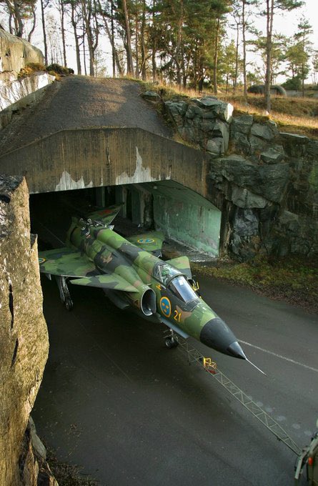 A camouflage-painted military fighter jet (numbered 27) emerging from a concrete bunker built into a rocky hillside surrounded by pine trees.
