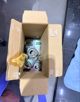 An open cardboard box filled with assorted aluminum beverage cans, viewed from above on a dark tiled floor.