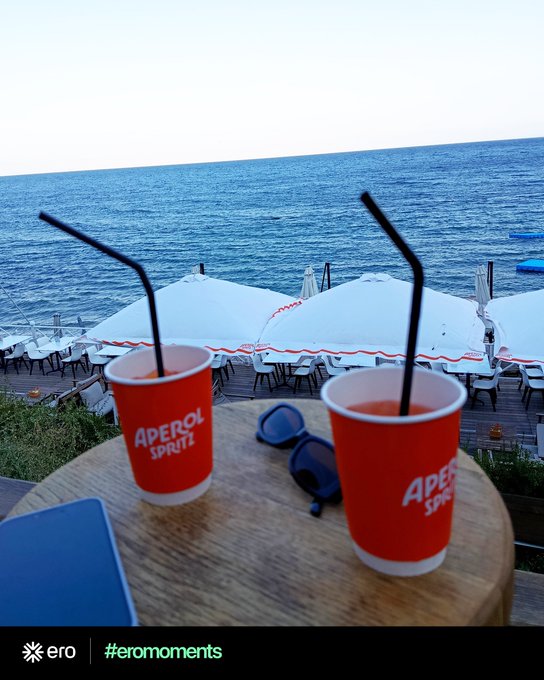 Two bright red "Aperol Spritz" cups with black bendy straws sit on a wooden table alongside sunglasses, overlooking a seaside terrace with white umbrellas and lounge chairs by the blue ocean—capturing a perfect aperitivo moment tied to the #eromoments campaign encouraging customers to share their ero purchases.