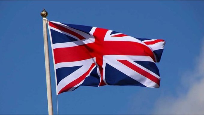 The image shows the United Kingdom's Union Jack flag waving against a clear blue sky. This connects to the post about the UK's Financial Conduct Authority (FCA) approving Bitcoin ETFs for retail customers, symbolizing the UK's regulatory milestone in the cryptocurrency market.