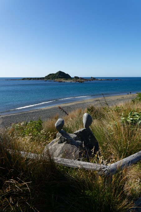 A coastal landscape featuring balanced stone sculptures in tall grass overlooking a pebble beach, calm blue ocean, and a small green island under a clear blue sky.