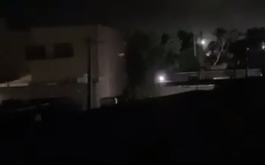 A dark, nighttime scene shows a building with bright lights in the distance, possibly indicating an explosion or fire, alongside trees and what appears to be damaged structures. The image's shadowy, tense atmosphere of destruction aligns with the post's description of Iran's southern region under drone attack, visually reinforcing the geopolitical instability contributing to BTC's retest of the 62.6k zone amid rising selling pressure.