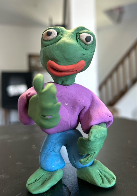 A handcrafted clay figurine of Pepe the Frog, painted green with a purple shirt and blue pants, displayed on a dark surface in a home setting.