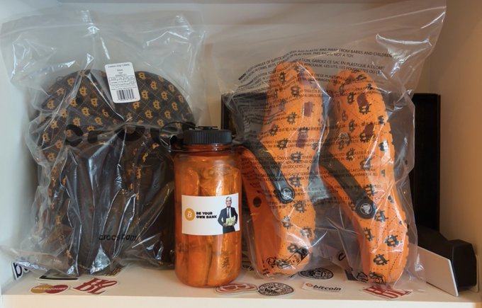 Two pairs of Crocs shoes in plastic packaging—one in the classic black/gold monogram pattern and one in orange with stars—alongside an orange Nalgene water bottle labeled 'BE YOUR OWN BOSS', all displayed on a Nike shoebox.
