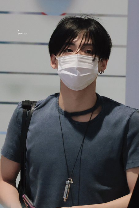 A young man wearing a white face mask and dark blue t-shirt, photographed indoors with a lanyard and strap visible.