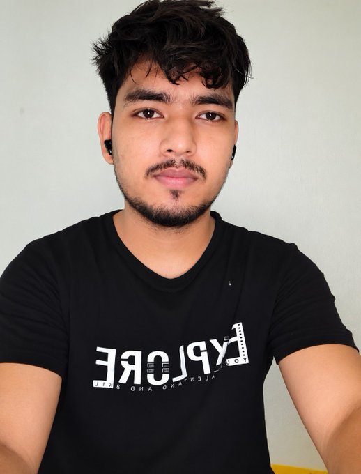 A young man with dark curly hair and a goatee wearing a black t-shirt with white 'EXPLORE THE WORLD' lettering, posing for a portrait against a plain wall.