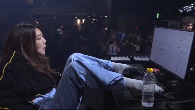 A young woman sits with her feet up on a gaming desk in front of a curved monitor displaying charts, with a water bottle beside her, in what appears to be an esports event setting.