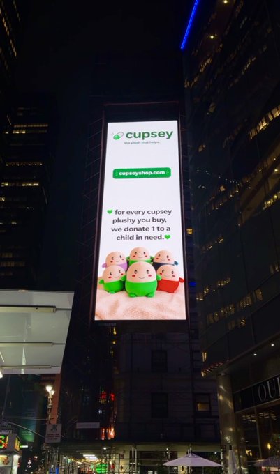 A large illuminated billboard in Manhattan advertises "Cupsey" plush toys, displaying the logo, website "cupseyshop.com," and the message "for every cupsey plushy you buy, we donate 1 to a child in need," featuring images of the cute green and pink plush characters, matching the post's announcement of Cupsey's presence in Manhattan.