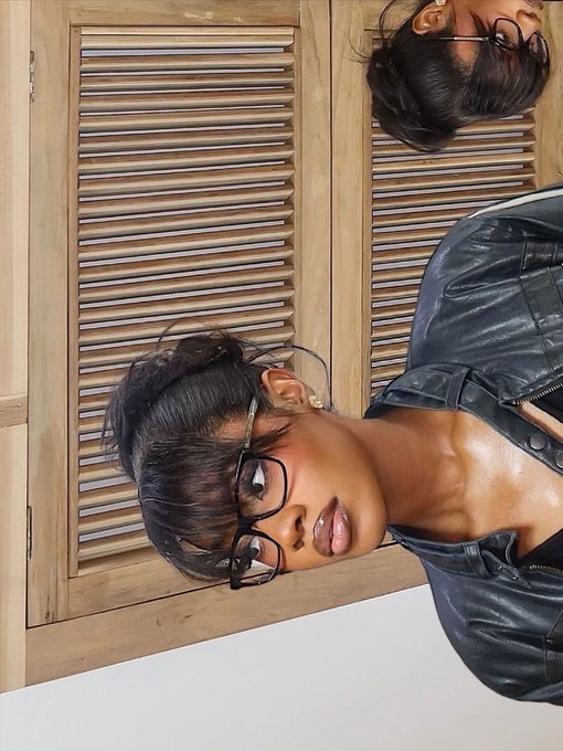 An upside-down selfie of a woman with short dark hair wearing glasses and a black leather jacket, posed against a wooden louvered door with her reflection visible.