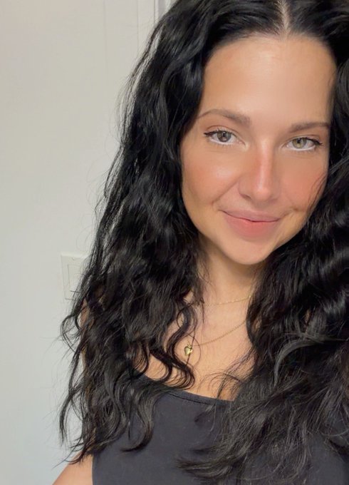 A selfie of a woman with long dark wavy hair, wearing a black top and a delicate gold necklace with small pendants.