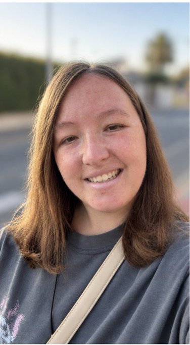 A smiling young woman with shoulder-length brown hair wearing a dark gray shirt and a white crossbody strap, photographed outdoors.