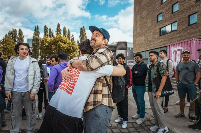 The image shows a group of people gathered together, with two men embracing each other in the center of the frame. The background includes a brick building and trees, creating an outdoor setting for this heartwarming moment. A caption below the photo reads "Capture @solana in 1 picture. I'll start." This suggests that the image is meant to be shared on social media platforms or used as part of a storytelling project.